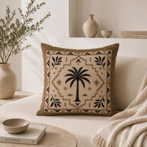 Peut inclure: Coussin carré avec une bordure marron et un centre beige orné d'un motif de palmier noir. Le coussin présente un motif géométrique et des accents de feuilles noires. Le coussin est sur un canapé blanc.