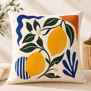 Peut inclure: Coussin carré avec un fond crème présentant un motif de citron coloré. Le motif comprend deux citrons jaunes, des feuilles vertes, des fleurs blanches et des formes abstraites bleues et orange. Le coussin est probablement en tissu doux.