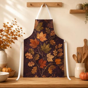 Peut inclure: Un tablier de cuisine présentant un fond marron foncé avec un motif répétitif de feuilles d'automne dans des tons orange, jaune, rouge et vert. Le tablier comprend des liens en tissu blanc au cou et à la taille pour le maintenir en place.