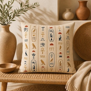 Peut inclure: Coussin carré beige avec un motif répété de hiéroglyphes égyptiens en bleu marine, rouge et or. Le coussin est posé sur un banc tressé avec un vase et des objets décoratifs en arrière-plan.
