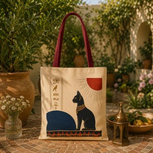 Peut inclure: Sac fourre-tout beige avec des anses bordeaux, orné d'un motif de chat égyptien. L'illustration comprend un chat noir, un soleil rouge et des symboles hiéroglyphiques. Le sac est en toile.
