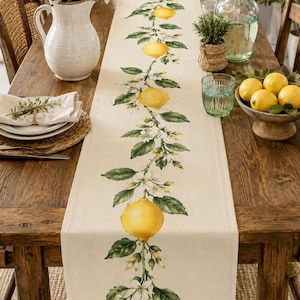 Peut inclure: Une nappe de table beige ornée d'un motif botanique de citron. La nappe présente des citrons jaunes, des feuilles vertes et des fleurs blanches. Elle est posée sur une table en bois avec d'autres éléments décoratifs.