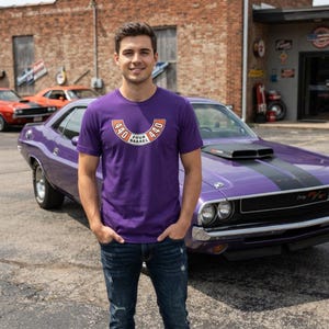 May include: A purple short-sleeve t-shirt featuring a graphic on the chest with the text 440 FOUR BARREL 440 in an orange and white semi-circular design. The shirt is paired with dark blue denim jeans with a distressed finish on the legs.