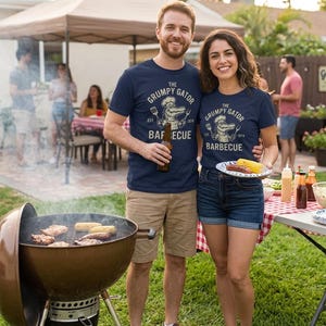 May include: A navy blue t-shirt featuring a graphic of an alligator wearing a chef hat, holding a spatula and fork, with the text "THE GRUMPY GATOR BARBECUE EST. 1974". The design is printed in a light cream color on the front of the shirt.
