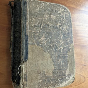 May include: An antique book with a distressed, weathered cover. The cover is a faded blue-gray with visible text and significant wear, including peeling and discoloration. The spine shows signs of age and use, with a visible binding thread.