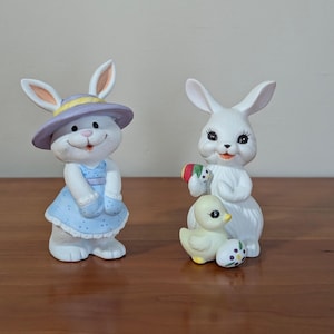 May include: Two ceramic Easter figurines. One is a white bunny wearing a blue dress and a lavender hat. The other is a white bunny holding a decorated egg and a yellow chick. The figurines are on a wooden surface.