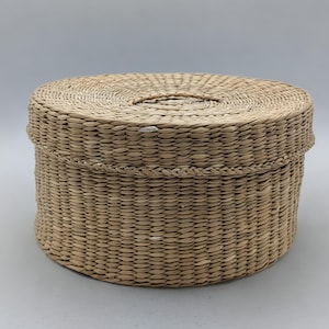 May include: A round, light tan woven basket with a matching lid. The basket features a tight, horizontal coil weave pattern made from natural dried plant fibers, creating a textured surface with a small, integrated handle on the lid.