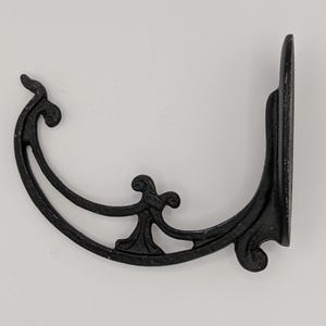 Könnte beinhalten: Schwarzer dekorativer Wandhalter aus Gusseisen mit einem verzierten, geschwungenen Design. Die Halterung weist Schnörkel auf und hat eine flache Rückseite zur Montage. Ideal zum Aufhängen von Pflanzen oder anderen Dekorationsgegenständen.