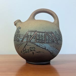 May include: A round, matte-finish ceramic pitcher with a top handle and a small spout. The surface features a muted, gradient-toned landscape illustration of a truss bridge spanning a river, rendered in dark lines against a tan and blue background.