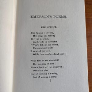 Puede incluir: P&aacute;gina de un libro abierto con el poema "THE SPHINX" de "EMERSON'S POEMS." El texto negro est&aacute; impreso en una p&aacute;gina crema. El n&uacute;mero "7" est&aacute; en la parte inferior de la p&aacute;gina. El libro est&aacute; sobre una superficie de madera.
