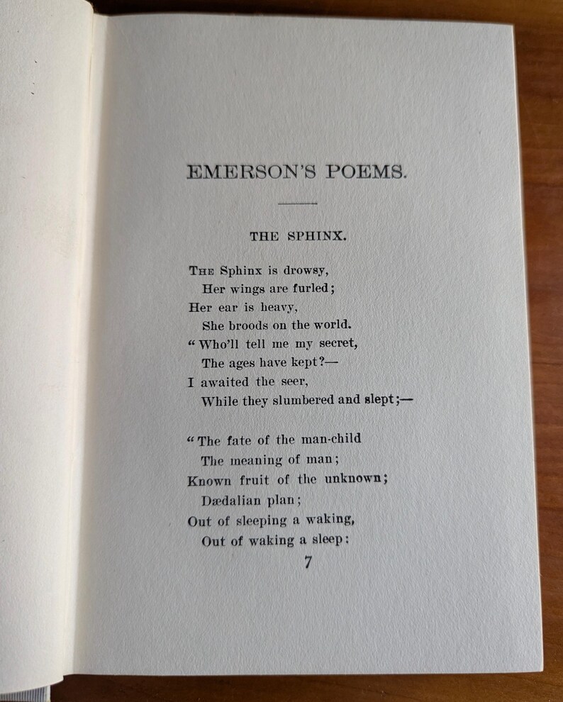 Puede incluir: P&aacute;gina de un libro abierto con el poema "THE SPHINX" de "EMERSON'S POEMS." El texto negro est&aacute; impreso en una p&aacute;gina crema. El n&uacute;mero "7" est&aacute; en la parte inferior de la p&aacute;gina. El libro est&aacute; sobre una superficie de madera.