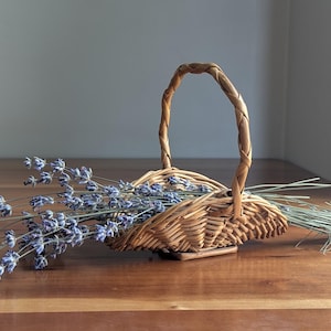 May include: A small, light brown woven wicker basket with a single arched handle, containing a bundle of dried lavender stems with purple flower buds and thin green stalks, arranged horizontally across the basket's opening.