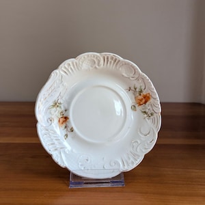 May include: A round, off-white ceramic saucer featuring an embossed, scalloped rim with intricate scrollwork and relief patterns. The center is smooth, and the border is decorated with delicate, hand-painted orange floral motifs and green leaves.