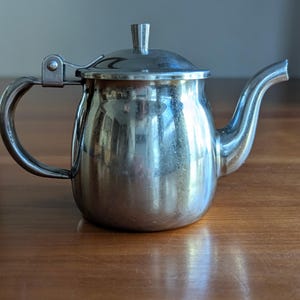 May include: A small, shiny silver teapot with a curved spout and a hinged lid. The handle is a simple, curved design. The teapot is sitting on a wooden surface.