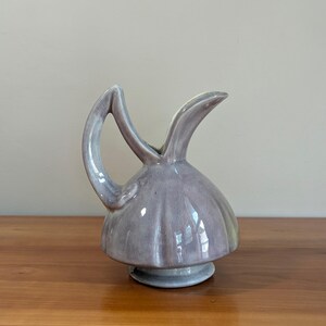 May include: A lavender-gray ceramic pitcher with a glossy finish, featuring a rounded, ribbed body, a pedestal base, and a high, curved handle that extends upward from the side to meet the top of the spout, creating a distinct silhouette.