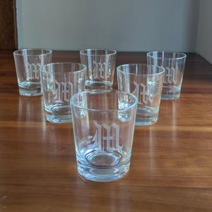 May include: Six clear glass tumblers with a frosted letter "M" etched on the front. The glasses are arranged on a dark wooden surface. The glasses are cylindrical and appear to be shot glasses or small drinking glasses.