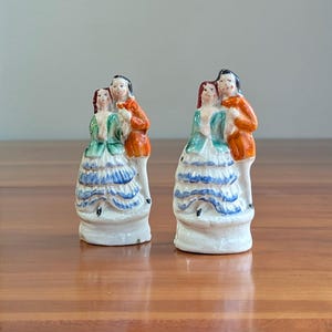 May include: Two ceramic figurines of a couple in period clothing. The woman wears a green and blue dress, while the man has an orange coat. The figurines are on a white base and are set on a wooden surface.