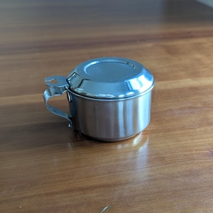 May include: A small, stainless steel container with a lid and a handle. The container is cylindrical with a rounded lid and a small handle on the side. The metal has a shiny, reflective surface. The container sits on a wooden surface.