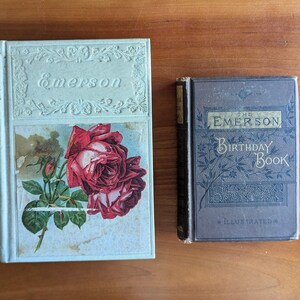 Puede incluir: Dos libros antiguos sobre una superficie de madera. El libro m&aacute;s grande es de color azul claro con el nombre "Emerson" en relieve en la portada y una ilustraci&oacute;n de rosa roja. El libro m&aacute;s peque&ntilde;o es azul oscuro con "The Emerson Birthday Book" en la portada.