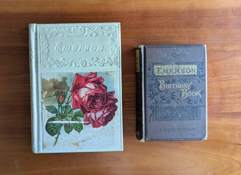 Puede incluir: Dos libros antiguos sobre una superficie de madera. El libro m&aacute;s grande es de color azul claro con el nombre "Emerson" en relieve en la portada y una ilustraci&oacute;n de rosa roja. El libro m&aacute;s peque&ntilde;o es azul oscuro con "The Emerson Birthday Book" en la portada.