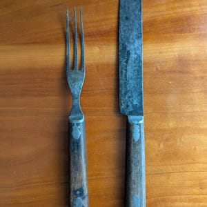 May include: A vintage fork and knife set with wooden handles and metal blades. The fork has three tines, and the knife has a rounded tip. The handles are dark brown, and the metal has a worn, antique appearance. The set is placed on a wooden surface.