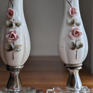May include: A pair of white porcelain candle holders featuring raised pink rose floral accents with green leaves. The stems are supported by silver-toned metal bases that connect to clear, square-shaped glass pedestals at the bottom.