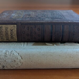 Puede incluir: Dos libros antiguos apilados. El libro superior, marr&oacute;n oscuro, tiene la inscripci&oacute;n dorada "The Emerson Birthday Book". El libro inferior es de color beige claro con dise&ntilde;os florales en relieve.