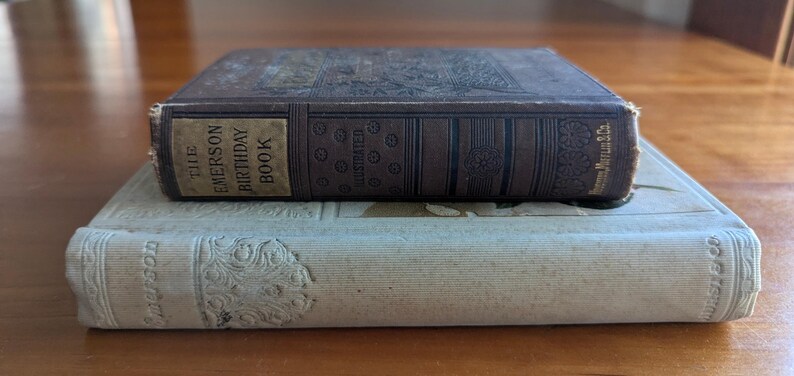 Puede incluir: Dos libros antiguos apilados. El libro superior, marr&oacute;n oscuro, tiene la inscripci&oacute;n dorada "The Emerson Birthday Book". El libro inferior es de color beige claro con dise&ntilde;os florales en relieve.