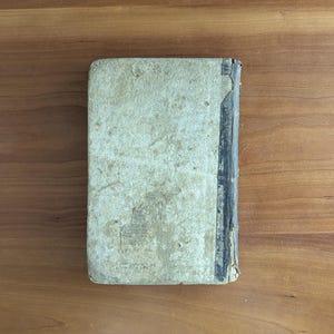 May include: A vintage book with a worn, off-white cover and a dark spine. The cover shows signs of age with discoloration and wear. The book is rectangular and appears to be closed, resting on a wooden surface.