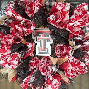 May include: A decorative wreath featuring a central Texas Tech University logo sign. The wreath is constructed with layers of black mesh, red and white plaid ribbon, black and white buffalo check ribbon, and tan burlap fabric accents.