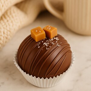 Könnte beinhalten: Eine Schokoladenkugel in einer weißen Papierform, belegt mit zwei Karamellwürfeln und Meersalz. Die Kugel hat diagonale Schokoladenlinien. Eine beige Tasse und eine beige Strickdecke befinden sich im Hintergrund.