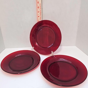May include: Three circular ruby red glass plates with smooth rims and a central recessed area. A vertical measuring tape is positioned behind the top plate, showing a diameter of approximately 20 cm for the glass dinnerware set.