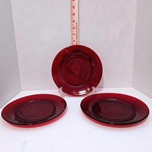 May include: Three circular plates made of deep red translucent glass. The plates feature a smooth, reflective surface and a slightly raised rim. A wooden ruler is positioned vertically behind the centre plate, showing a measurement of 15 cm.
