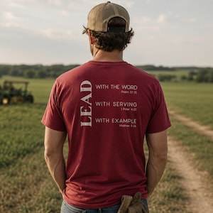 May include: A maroon short-sleeved t-shirt featuring white text on the back that reads: LEAD WITH THE WORD Psalm 37:31, WITH SERVING 1 Peter 4:10, WITH EXAMPLE Matthew 5:16. The shirt is worn with a tan baseball cap and blue denim jeans.