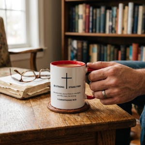 May include: A white ceramic mug with a red interior and handle, featuring a black cross graphic and the text: BE STRONG STAND FIRM. Be watchful, stand firm in the faith, act like men, be strong. 1 CORINTHIANS 16:13. The mug sits on a brown coaster.