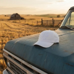 May include: A white baseball cap featuring the embroidered text Psalm 34:14 on the front panel. The cap has a curved brim, a structured crown with a button top, and tonal stitching throughout the fabric, resting on a weathered blue metal surface.