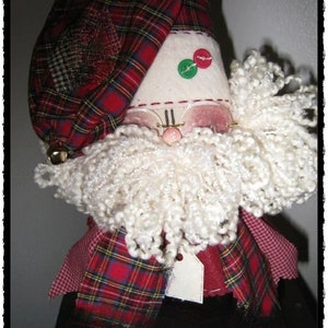 Pode incluir: Um boneco de Papai Noel feito à mão com um chapéu xadrez vermelho e verde, barba branca e óculos. O boneco está usando uma echarpe xadrez vermelha e verde e tem uma etiqueta branca pendurada no pescoço.