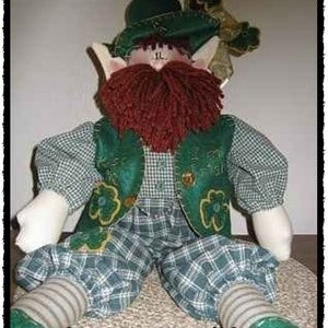 Puede incluir: Un muñeco leprechaun de cuadros verdes y blancos con una barba de lana roja, un sombrero verde y zapatos verdes. El muñeco está sentado sobre una superficie marrón y tiene el texto "Kiss Me, I'm Irish E-pattern" en la parte inferior de la imagen.
