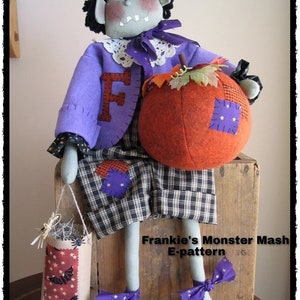 Könnte beinhalten: Eine handgemachte Halloween-Monsterpuppe mit lila Kleidung, einem karierten Rock und einem Kürbis. Die Puppe sitzt auf einer Holzkiste mit einer Tüte Süßigkeiten in der Hand. Der Text "Frankie's Monster Mash E-pattern" ist auf der Kiste zu sehen.