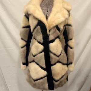 May include: A vintage fur coat with a cream-colored collar and geometric pattern in shades of cream, gray, and black. The coat is displayed on a mannequin, showcasing its design and texture. The coat has a black velvet stripe down the front.