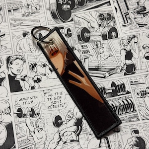 以下が含まれることがあります： 黒い長方形のキーホルダー、シルバーのリング付き。キーホルダーには、白い髪と赤い目のキャラクターのマンガ風イラストが描かれています。背景は、重量挙げをテーマにした白黒の漫画パネルです。