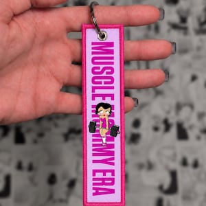 Könnte beinhalten: Ein rechteckiger Schlüsselanhänger aus Stoff mit einem leuchtend pinken gestickten Rand und einer hellvioletten Mitte. Der Text "MUSCLE MOMMY ERA" ist in fetten pinken Buchstaben aufgedruckt, ergänzt durch eine Comicfigur mit Hantel.