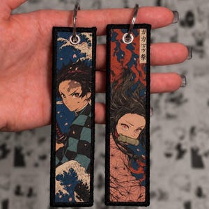 Pode incluir: Dois porta-chaves retangulares de tecido com bordas costuradas em preto e argolas de metal. Um apresenta uma personagem com haori xadrez verde e preto sobre um fundo de ondas azuis. O outro mostra uma personagem com um bocal de bambu e detalhes vermelhos.