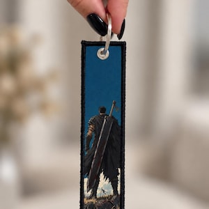 Peut inclure: Un porte-clés rectangulaire en tissu avec une illustration imprimée d'un guerrier portant une grande épée sur un champ de bataille. Le porte-clés présente une bordure noire cousue, un œillet métallique argenté et un anneau fendu argenté.