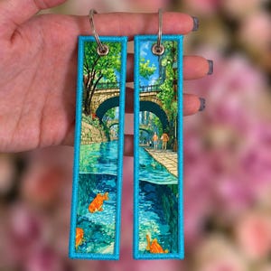 Pode incluir: Dois porta-chaves retangulares bordados com uma cena vibrante de uma ponte de pedra sobre um canal com árvores verdes exuberantes e peixes koi laranja a nadar na água. Cada porta-chaves tem uma borda azul brilhante costurada.
