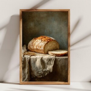 May include: A framed print of a still life featuring a loaf of bread and a slice on a draped table. The bread has a golden-brown crust and a light interior. A sprig of wheat is also on the table. The background is a muted, dark green.
