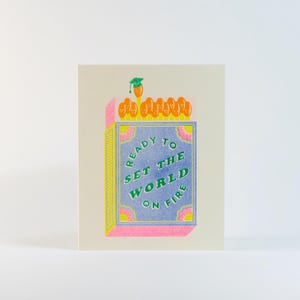 Może przedstawiać: Kartka okolicznościowa z motywem pudełka zapałek. Pudełko jest niebieskie z napisem "READY TO SET THE WORLD ON FIRE" w kolorze zielonym i żółtym. Zapałki są pomarańczowe z zieloną czapką absolwenta. Karta ma różową i żółtą obwódkę.