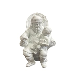 May include: A matte white ceramic figurine depicting Santa Claus seated in a high-backed chair with a small child on his lap. Santa wears a textured hat and coat, while the child holds a small gift. The entire piece is monochromatic and detailed.