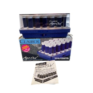 May include: A Clairol Aqua-Curl steam hairsetter with a blue base and clear lid, containing multiple dark blue and white hair rollers. The set includes the original cardboard packaging with product text and a printed instruction manual.