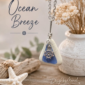 Puede incluir: Un collar de cadena plateada con un colgante triangular de vidrio marino blanco y azul, adornado con un pequeño dije de timón de barco plateado. El texto Ocean Breeze y Design by Mariel son visibles en la imagen.